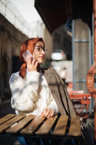 Eine Frau mit Hijab und Brille spricht an einem sonnigen Außentisch am Telefon.