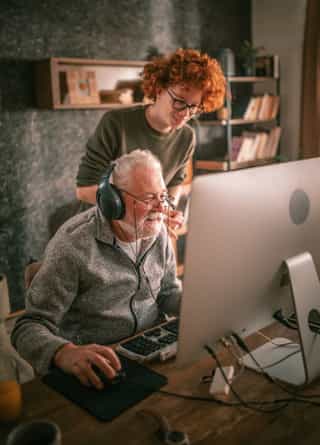 An older person with headphones sits at a computer desk, while a younger person with curly red hair stands behind, both focusing on the screen.