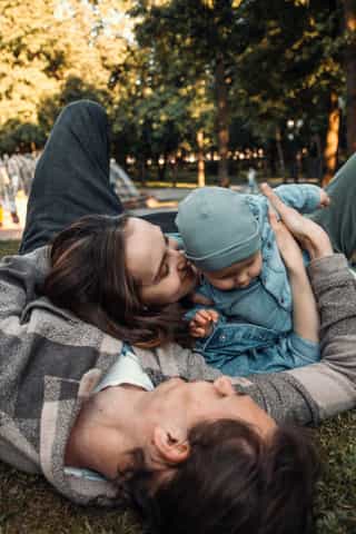 A baby with a blue cap and jeans clothing lies on a parent lying in the grass. The adult kisses the cheek of the baby and both enjoy a warm, sunny day in the park with trees in the background.